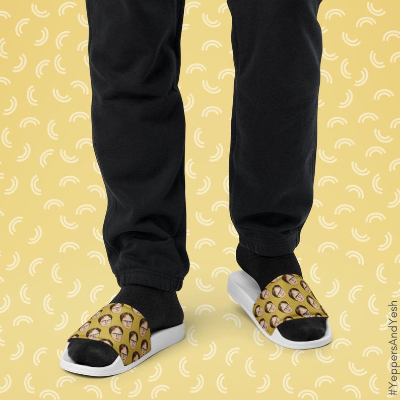 May include: White slide sandals featuring a repeating pattern of a person's face on the top strap. The background of the strap is a mustard yellow color. The sandals are worn with black socks and black sweatpants.