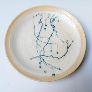 May include: A round ceramic plate with a cream-colored center and a light tan rim. The center features a dark blue, hand-painted botanical design depicting thin, branching stems with small, round berries scattered across the surface.