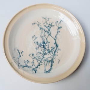 Op de afbeelding: Een ronde keramische bord met een glanzend crèmekleurig glazuur en een opstaande rand. Het midden van het bord is voorzien van een gedetailleerde, donkerblauwe botanische print van een vertakte boom of struik met kleine knoppen.