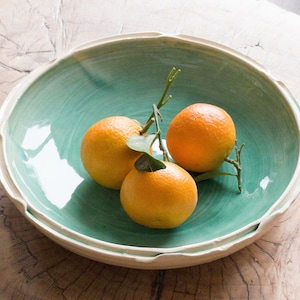 May include: Three oranges in a green ceramic bowl with a white rim. The oranges are arranged in a cluster with a sprig of green leaves.