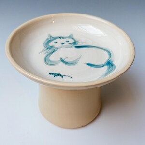 Puede incluir: Un cuenco de cerámica color crema sobre pedestal con una ilustración pintada a mano en azul de un gato tumbado junto a un pequeño ratón. El cuenco presenta una base amplia y poco profunda, además de un soporte cilíndrico firme y liso.