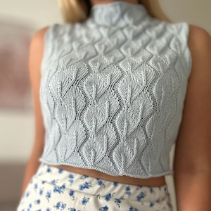 Könnte beinhalten: Ein hellblaues ärmelloses Strick-Crop-Top mit einem Blatt-Muster. Das Oberteil hat einen Stehkragen und eine figurbetonte Silhouette.