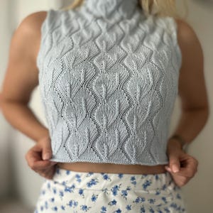 Könnte beinhalten: Ein hellblaues ärmelloses Stricktop mit Stehkragen und einem Zopfmuster. Das Top ist kurz und hat eine figurbetonte Silhouette.