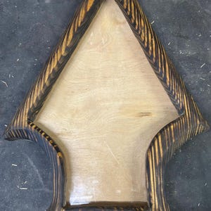 May include: A wooden arrow-shaped display shelf with a light wood interior and a dark, striped, burnt-wood frame. The shelf has a unique, handcrafted design, perfect for displaying small items. The arrow shape adds a rustic, artistic touch.
