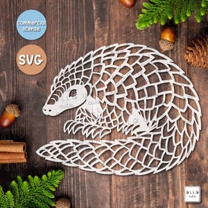 May include: A white cutout design of a pangolin on a brown wooden background. The design is intricate and detailed, with scales covering the pangolin's body. The design is available in SVG, PDF, PNG, and EPS formats.