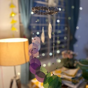 Peut inclure: Mobile suspendu avec un élément circulaire tressé et des éléments irisés en forme de feuilles violettes et vertes. Le mobile est suspendu devant une fenêtre avec des guirlandes lumineuses et une lampe allumée.