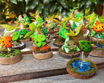Puede incluir: Una colección de figuritas de arcilla en miniatura que representan diversas criaturas del bosque, cada una con un diseño único y pintada en colores vibrantes. Las figuritas se exhiben en pequeñas bases de madera con musgo y otros elementos naturales.