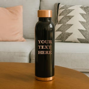 May include: A black insulated water bottle with a copper-colored lid and base. The words "YOUR TEXT HERE" are printed in a rose gold color on the front of the bottle. The bottle is sitting on a wooden table.