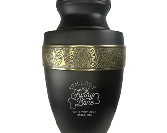 May include: Black urn with a gold band and engraved text that reads "Still got my funny bone". The urn has a decorative owl design on the band.