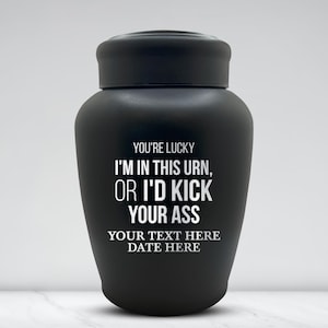 May include: A matte black urn with a lid. The urn has white text that reads "YOU'RE LUCKY I'M IN THIS URN, OR I'D KICK YOUR ASS". Below the text are the words "YOUR TEXT HERE DATE HERE".