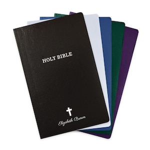 May include: A stack of six personalized Holy Bibles in different colors, including black, white, blue, green, and purple. The black Bible has the words "HOLY BIBLE" and a cross with the name "Elizabeth Brown" printed on the front cover.