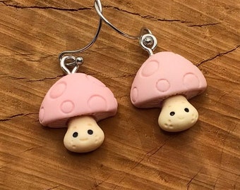 May include: A pair of pink mushroom earrings with white smiling faces. The earrings are made of polymer clay and have a silver hook.