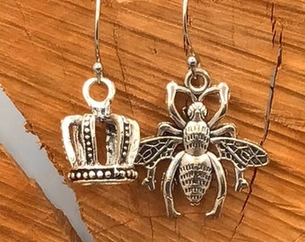 Puede incluir: Un par de pendientes de plata, uno con forma de corona y el otro con forma de abeja.