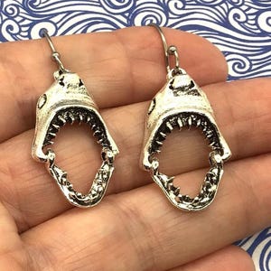 May include: Silver shark earrings with open mouths and sharp teeth. The earrings are hanging from a person's ear.