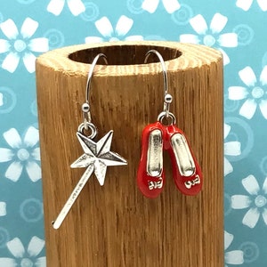 May include: A pair of silver dangle earrings. One earring features a silver star-shaped wand, and the other earring features a pair of red ruby slippers.