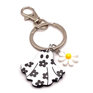 May include: A silver keychain with a ghost-shaped charm decorated with black flowers and a white daisy charm with a yellow center. The keychain has a lobster clasp and a split ring.