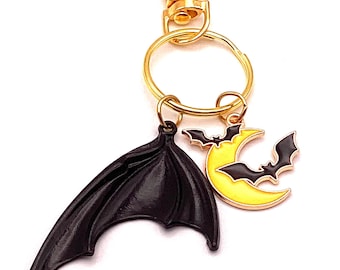May include: A gold-colored keychain with a lobster clasp, a key ring, and three charms. The charms include a black bat wing, a yellow crescent moon with two black bats, and a black bat.