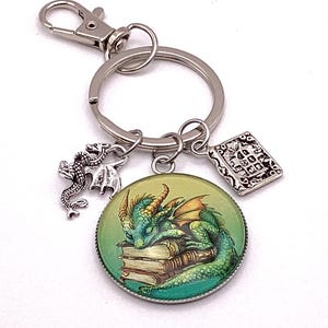 May include: A silver-tone keychain with a round charm featuring a green dragon sleeping on books. Additional charms include a small dragon and a book. The keychain has a lobster clasp and a split ring.