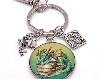 May include: A silver-tone keychain with a round charm featuring a green dragon sleeping on books. Additional charms include a small dragon and a book. The keychain has a lobster clasp and a split ring.