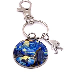 May include: A Bigfoot-themed keychain featuring a round charm with a starry night design and a silver-colored Bigfoot charm. The round charm depicts a Bigfoot figure standing near a lake under a starry sky. The keychain has a silver clasp and a keyring.