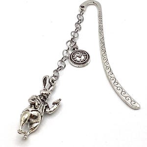 May include: A silver-toned metal bookmark featuring a charm of the White Rabbit from Alice in Wonderland, holding a pocket watch. The bookmark has a decorative swirl design and a chain with a small clock charm.