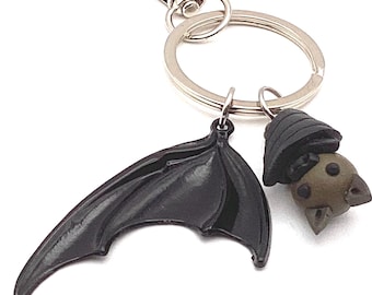 May include: A keychain featuring a black bat wing and a small bat figure with a dark hat. The keychain has a silver clasp and a ring for keys. The bat figure is brown with dark spots.