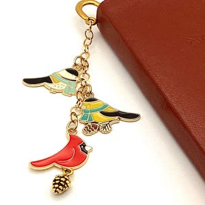 May include: A gold-toned keychain with three bird charms. The charms include a red cardinal with a pinecone, and two birds with black, white, yellow, and blue details. The keychain is attached to a brown leather item.