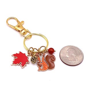 Puede incluir: Un llavero dorado con un mosquet&oacute;n y un anillo partido. Los dijes incluyen una hoja de arce roja, una peque&ntilde;a pi&ntilde;a y una ardilla con esmalte naranja y marr&oacute;n. Tambi&eacute;n hay una cuenta roja y una moneda.