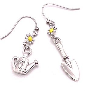 May include: Silver-toned garden-themed earrings. One earring features a watering can charm with a flower design, the other a trowel. Each earring has a daisy with yellow centers. The earrings are on a white background.