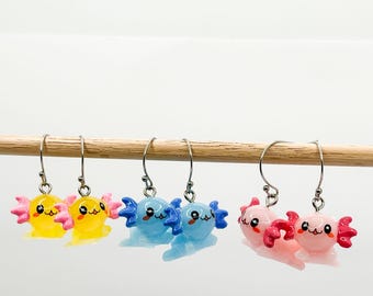 Peut inclure: Six paires de boucles d'oreilles axolotl, deux paires sont jaunes, deux paires sont bleues et deux paires sont roses. Les boucles d'oreilles sont suspendues à une baguette en bois.