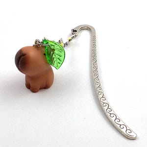 May include: A whimsical bookmark featuring a brown capybara figurine with a green leaf accent. The bookmark has a silver metal hook with a decorative swirl design. A charming accessory for book lovers.