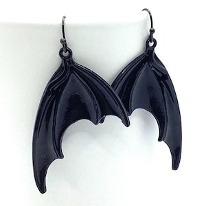 May include: A pair of black dangle earrings featuring a bat wing design. The wings are detailed with a textured surface and have a slightly curved shape.