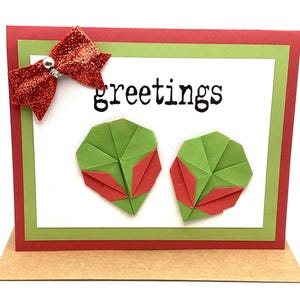 May include: A handmade greeting card with a red and green color scheme. The card features the word "greetings" in black text, two origami leaf designs, and a sparkly red bow. The card is displayed on a brown stand.