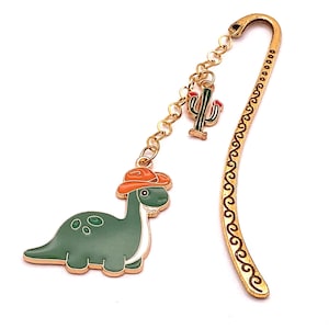 May include: A gold-colored metal bookmark with a green dinosaur wearing an orange cowboy hat. A small cactus charm and a decorative swirl design are also present. The bookmark is designed for books and reading.