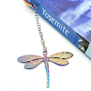 May include: A rainbow iridescent dragonfly bookmark with a silver chain attached to a metal clip. The bookmark is hanging from a book titled "Yosemite".