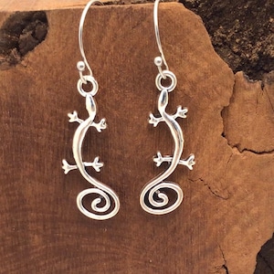 May include: A pair of silver-colored lizard earrings. Each earring features a lizard design with a curled tail. The earrings are suspended from a hook. The background is a textured, brown surface.