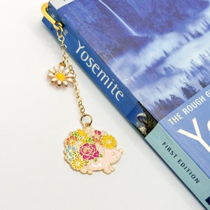 May include: Gold bookmark featuring a pink hedgehog with colorful flowers. The bookmark is attached to a chain with a white daisy charm.