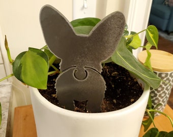 May include: A metal silhouette of a dog's rear end, sticking out of a white pot with green leafy plants. The dog silhouette is gray and has a tail.