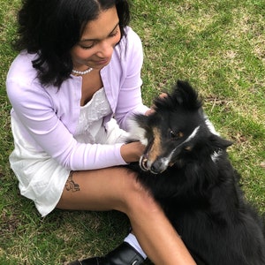 Könnte beinhalten: Eine junge Person in einem helllila Cardigan und einem weißen Kleid sitzt auf dem Gras mit einem schwarz-weißen Hund. Die Person trägt schwarze Stiefel und eine Perlenkette.
