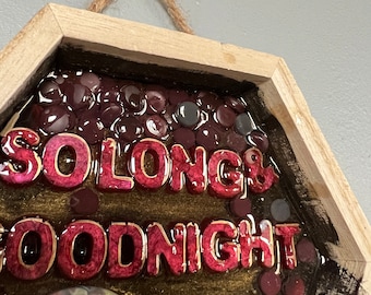 May include: A hexagonal wooden frame with a resin art piece inside. The resin art features a black background with gold glitter, red and black beads, and a pink and white swirl. The words "So Long & Goodnight" are written in pink letters with gold outlines.