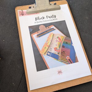 May include: A wooden clipboard holds a white sheet with the title "Block Party: A Quilty Colouring Book." The image features a coloring book, colored pencils, and a small package of colored pencils. The clipboard has a metal clip at the top.