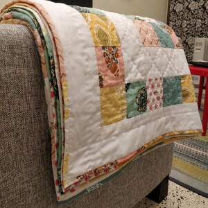 May include: A white quilt with a patchwork design featuring squares of pink, yellow, green, and blue fabric with floral patterns. The quilt is folded and draped over a grey sofa.
