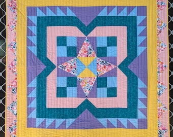 May include: A quilt with a star-shaped design in the center. The quilt is made of various fabrics in shades of pink, purple, teal, and yellow. The quilt has a scalloped border with a floral pattern.
