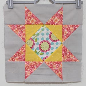 Puede incluir: Un bloque de colcha gris y blanco con un patrón floral rosa y blanco en el centro. El bloque está formado por ocho triángulos y un cuadrado en el centro. Los triángulos están dispuestos en forma de estrella.