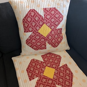 May include: Two decorative throw pillows with a red and yellow floral design on a white background. The pillows are on a black sofa.