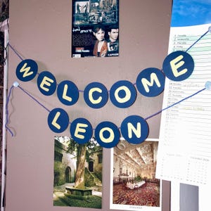 May include: A decorative banner featuring dark blue circular cardstock cutouts strung together with purple yarn. Each circle displays a single pale yellow letter, collectively spelling out the phrase WELCOME LEON in two distinct rows.