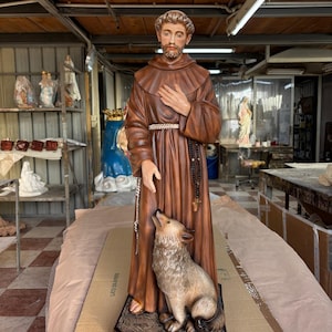 Op de afbeelding: Gedetailleerd beeld van een religieuze figuur in een bruin gewaad, met een wolf aan zijn voeten. Het beeld heeft een serene uitdrukking en houdt zijn hand op zijn borst. De wolf is lichtbruin en lijkt naar de figuur op te kijken.