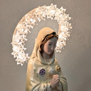 Peut inclure: Figurine religieuse d'une femme en robe cr&egrave;me, avec un halo de lumi&egrave;res en forme d'&eacute;toiles. La figurine a une expression sereine et est orn&eacute;e d'ornements en forme de roses. Un chapelet est suspendu au cou de la figurine.