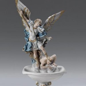 Könnte beinhalten: Ein weißer Keramik-Weihwasserfont mit einer Skulptur eines Engels, der einen Dämon besiegt. Der Engel ist mit Flügeln, einem Schwert und einem blauen und goldenen Gewand dargestellt. Der Dämon liegt unter dem Fuß des Engels.