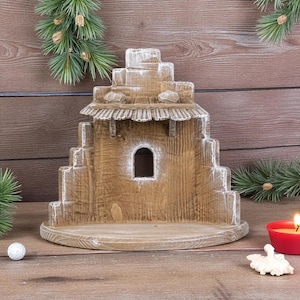 Può includere: Un presepe in legno con un design a gradini, con una piccola finestra ad arco e un tetto strutturato. La struttura è dipinta con accenti bianchi e poggia su una base rotonda. La scena è ambientata su una superficie di legno.